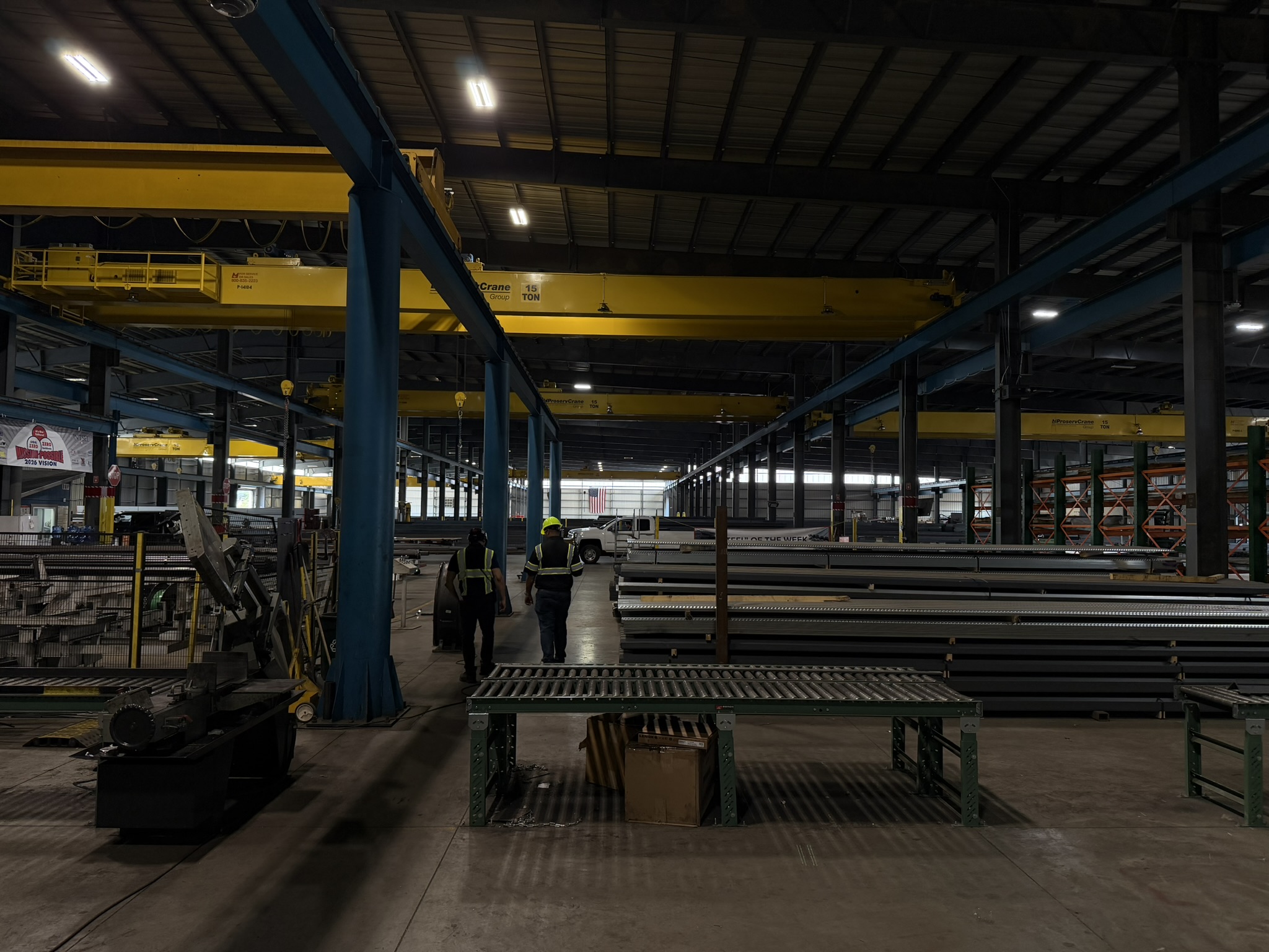 Workers walk through an American manufacturing facility beneath overhead cranes and an American flag.
